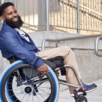 Wes doing a wheely in his wheelchair wearing a blue sport coat and khaki pants provided by Tommy Hilfiger for the IMIXX adaptive fashion event.